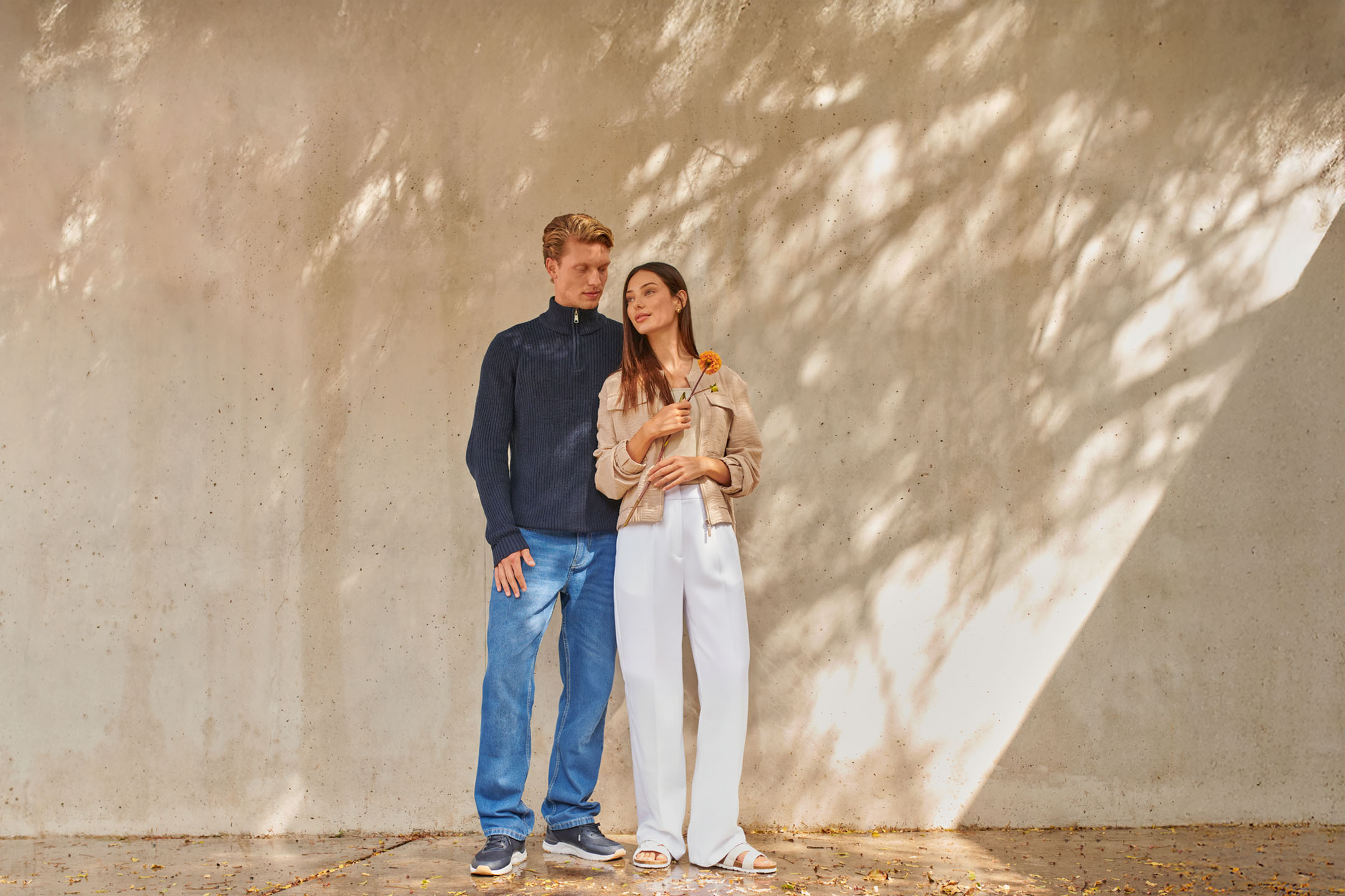 Man in sweater and jeans, woman in jacket and wide-leg trousers, holding a flower.