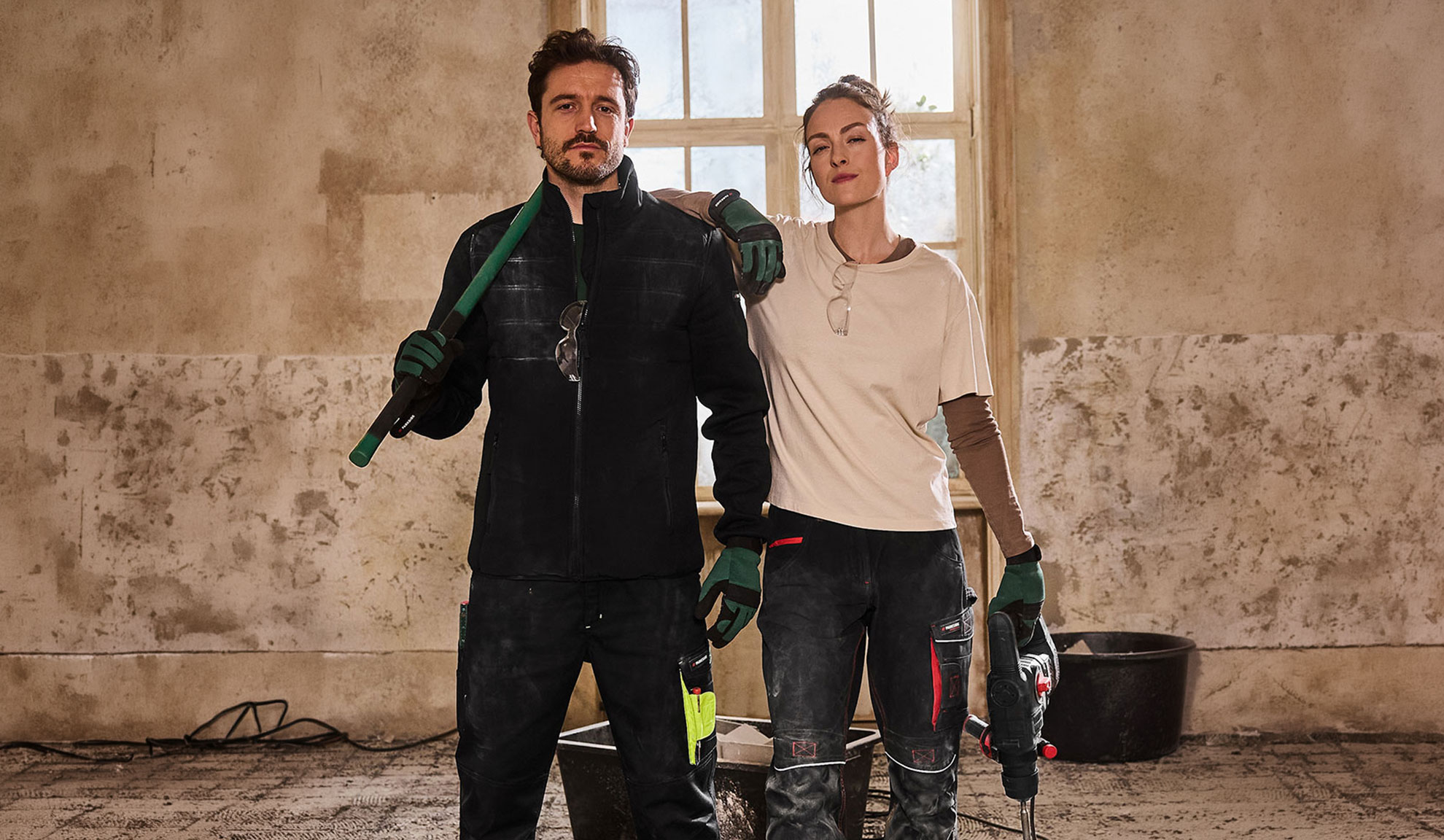Two construction workers in workwear, man with shovel and woman with drill.