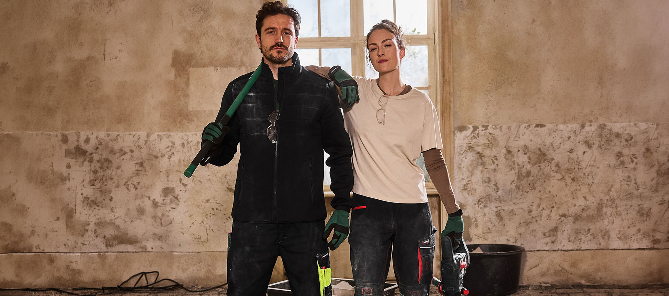 Man and woman in workwear, holding tools, standing in a room under renovation.