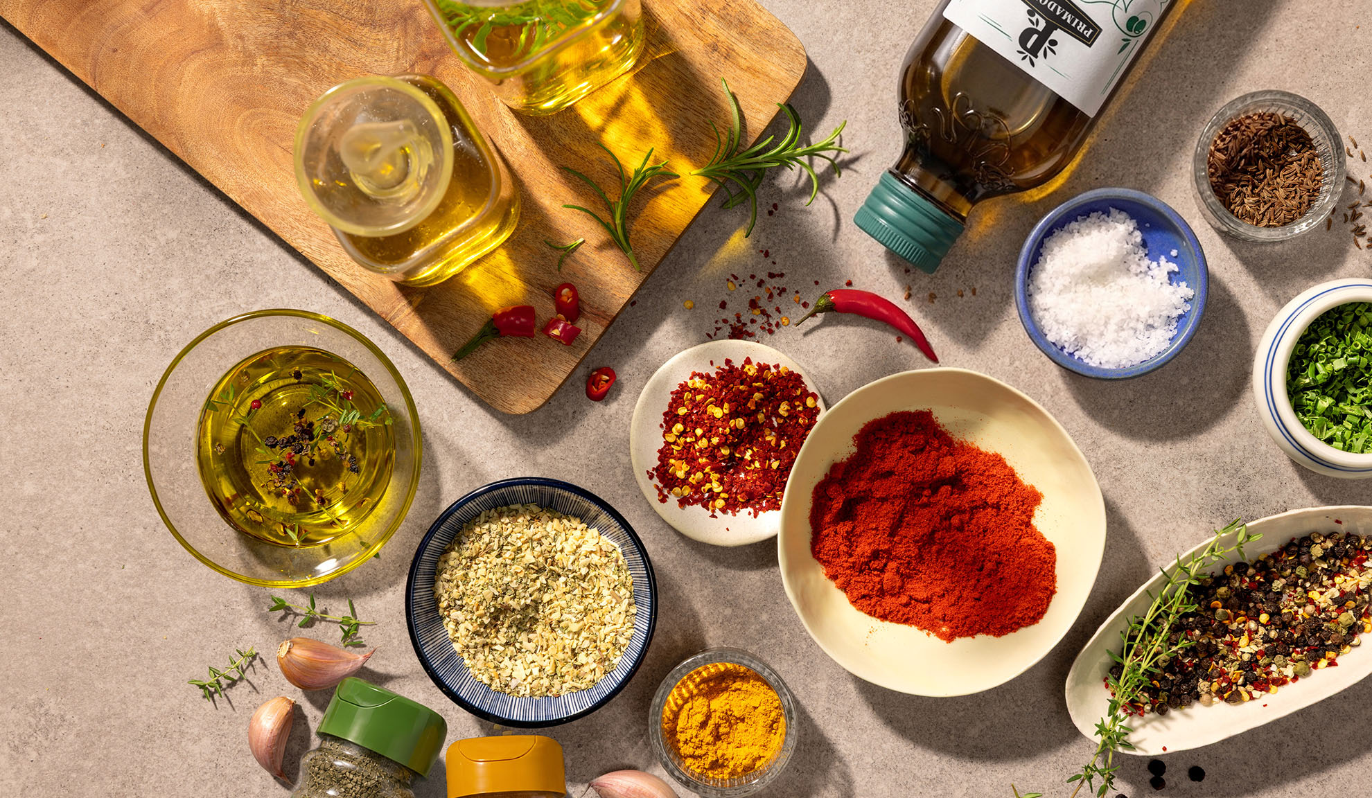 Olive oil, spices, and chilies on a wooden cutting board and concrete countertop.