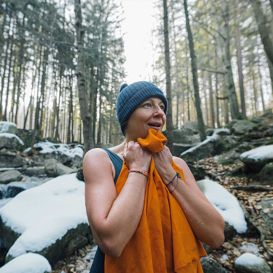 Woman in a beanie and swimsuit warming up with a towel in a snowy forest.