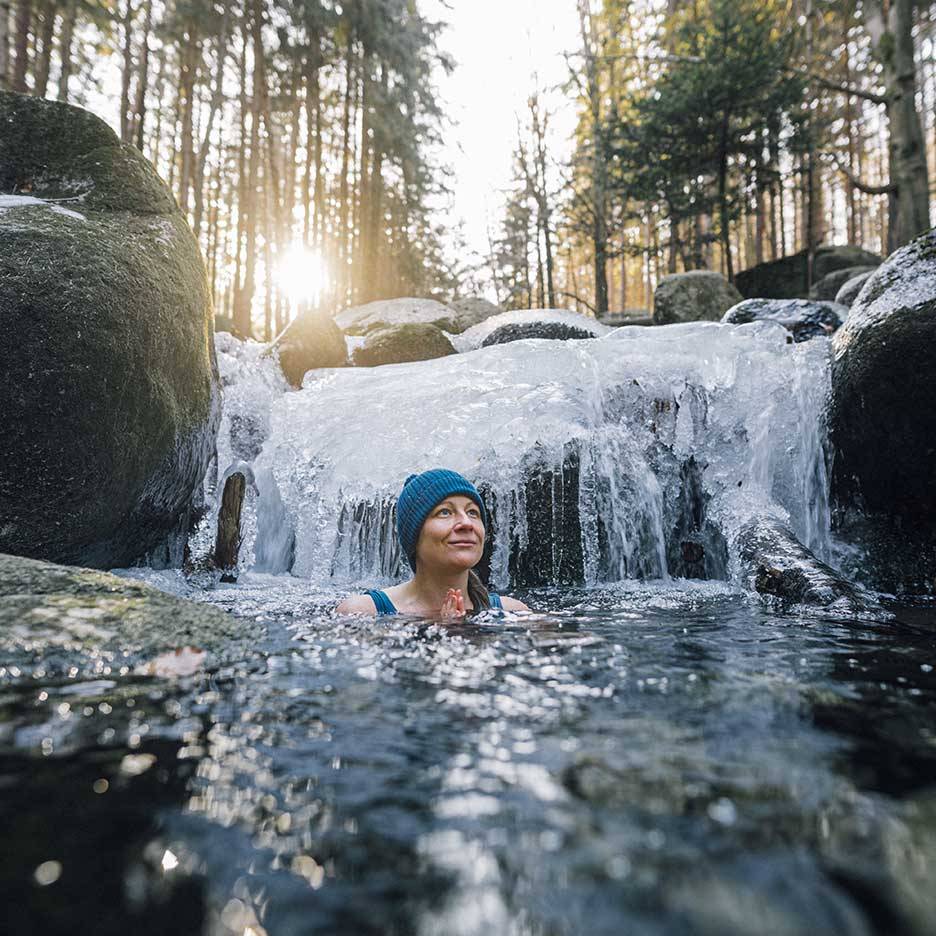 Woman in a blue hat and swimsuit enjoying a cold plunge in a frozen river.