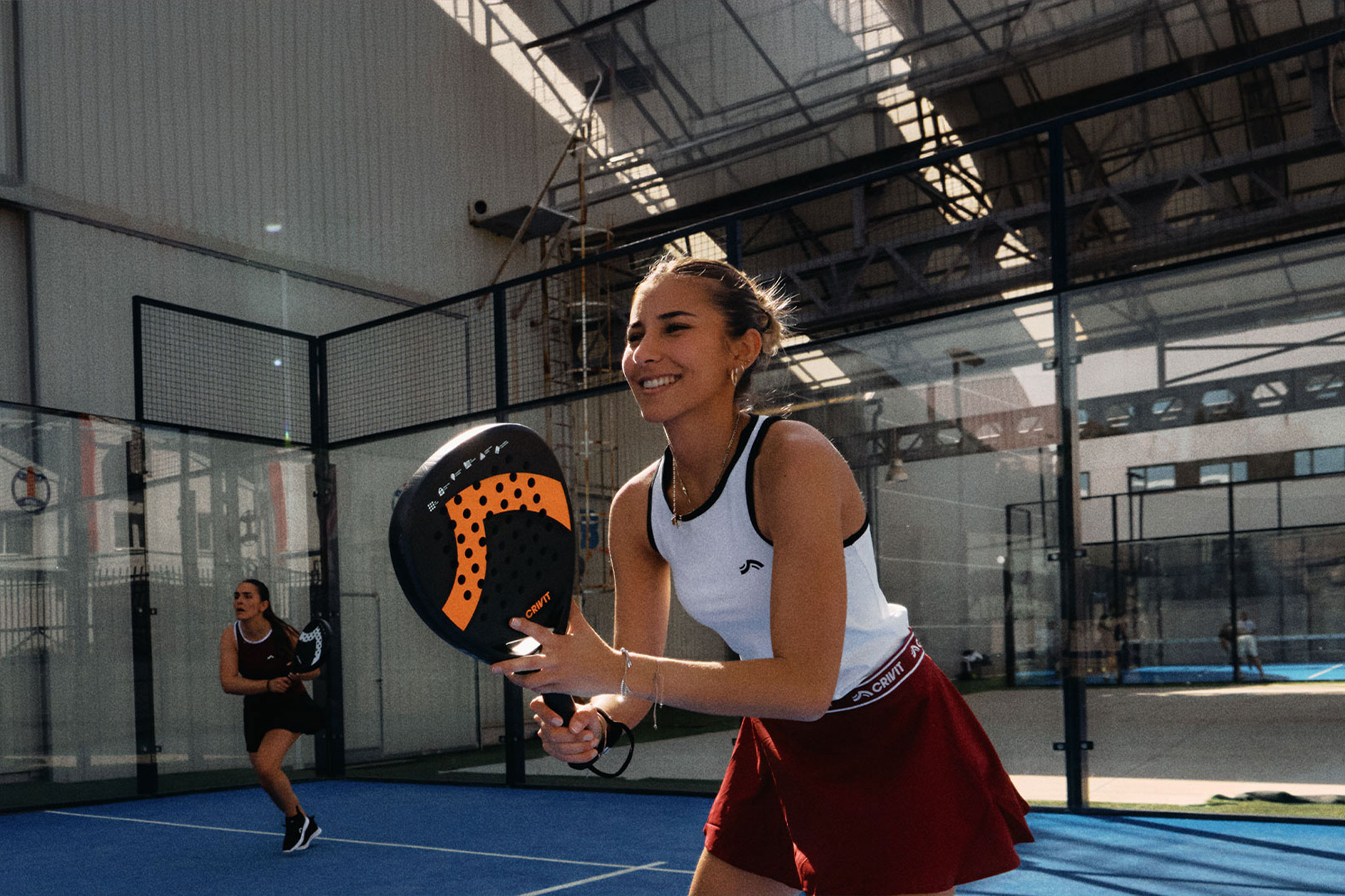 undefined: Two women playing padel tennis, one smiling and holding a racket, on a blue court.