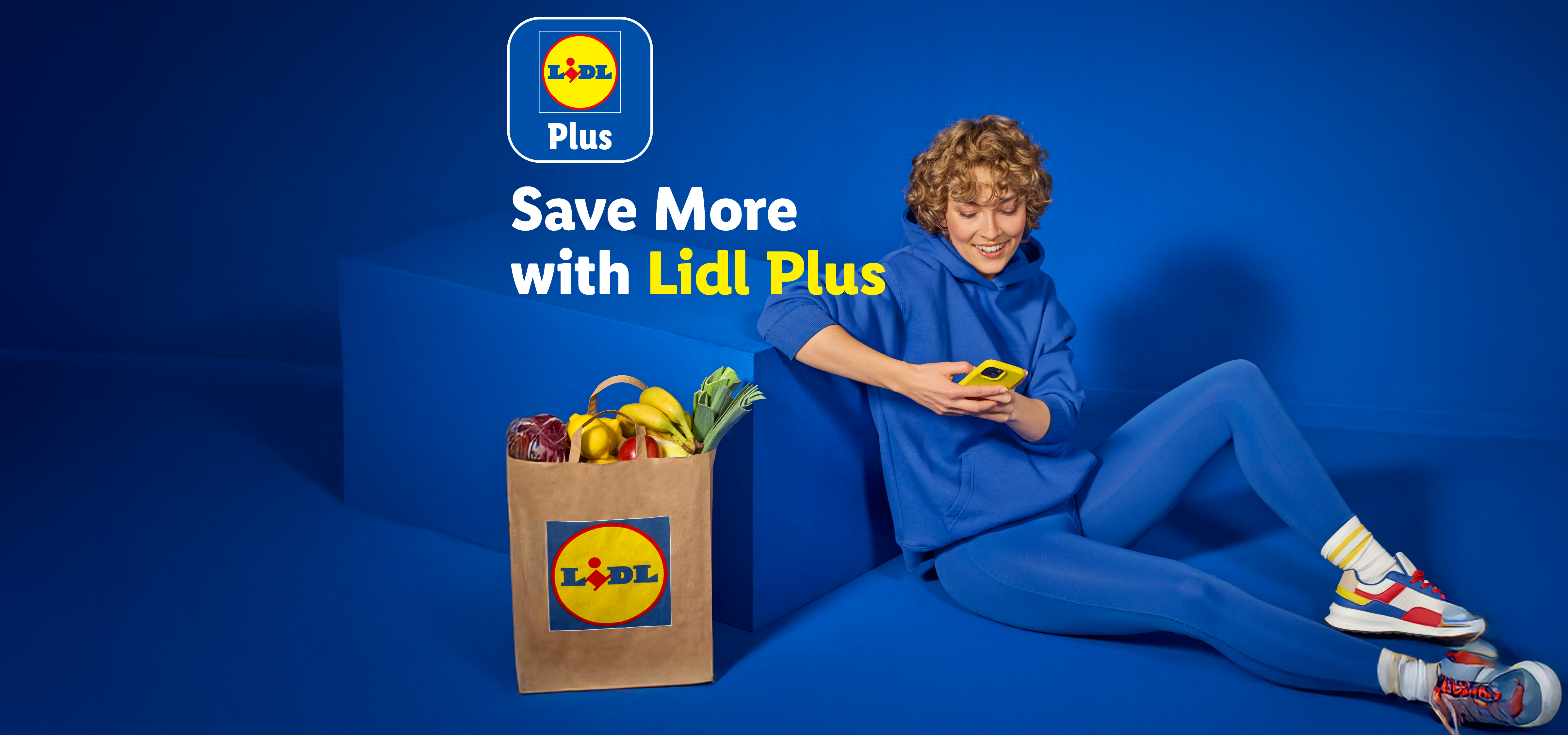 A person using a phone next to a grocery bag with fruits and vegetables, with text 'Save More with Plus'.