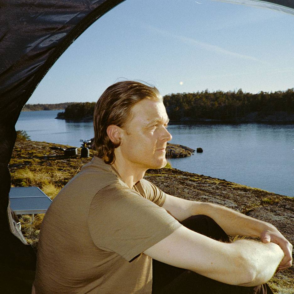 Man in a sports t-shirt sitting in a tent, looking out at a lake and forest.