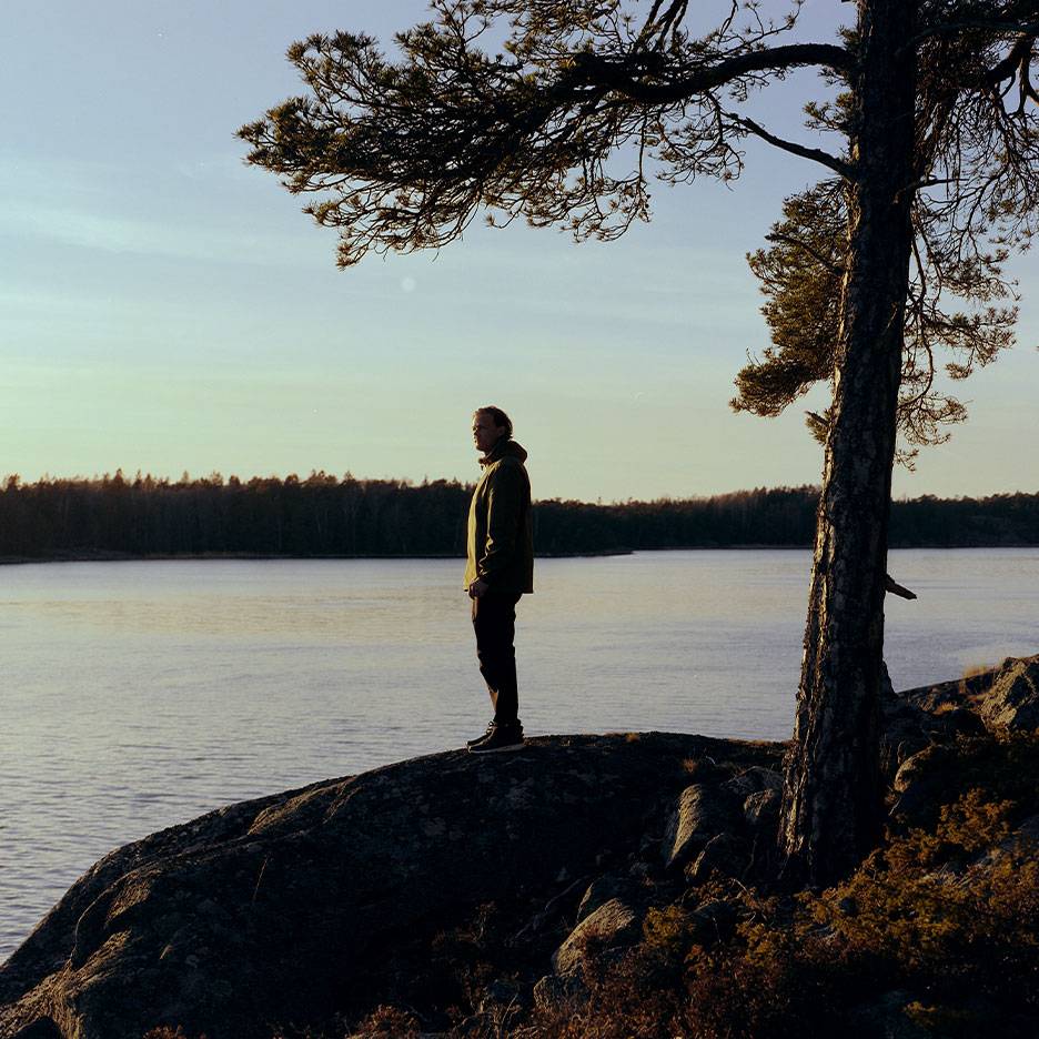 Man in a hiking jacket by a lake, with a tree in the foreground.