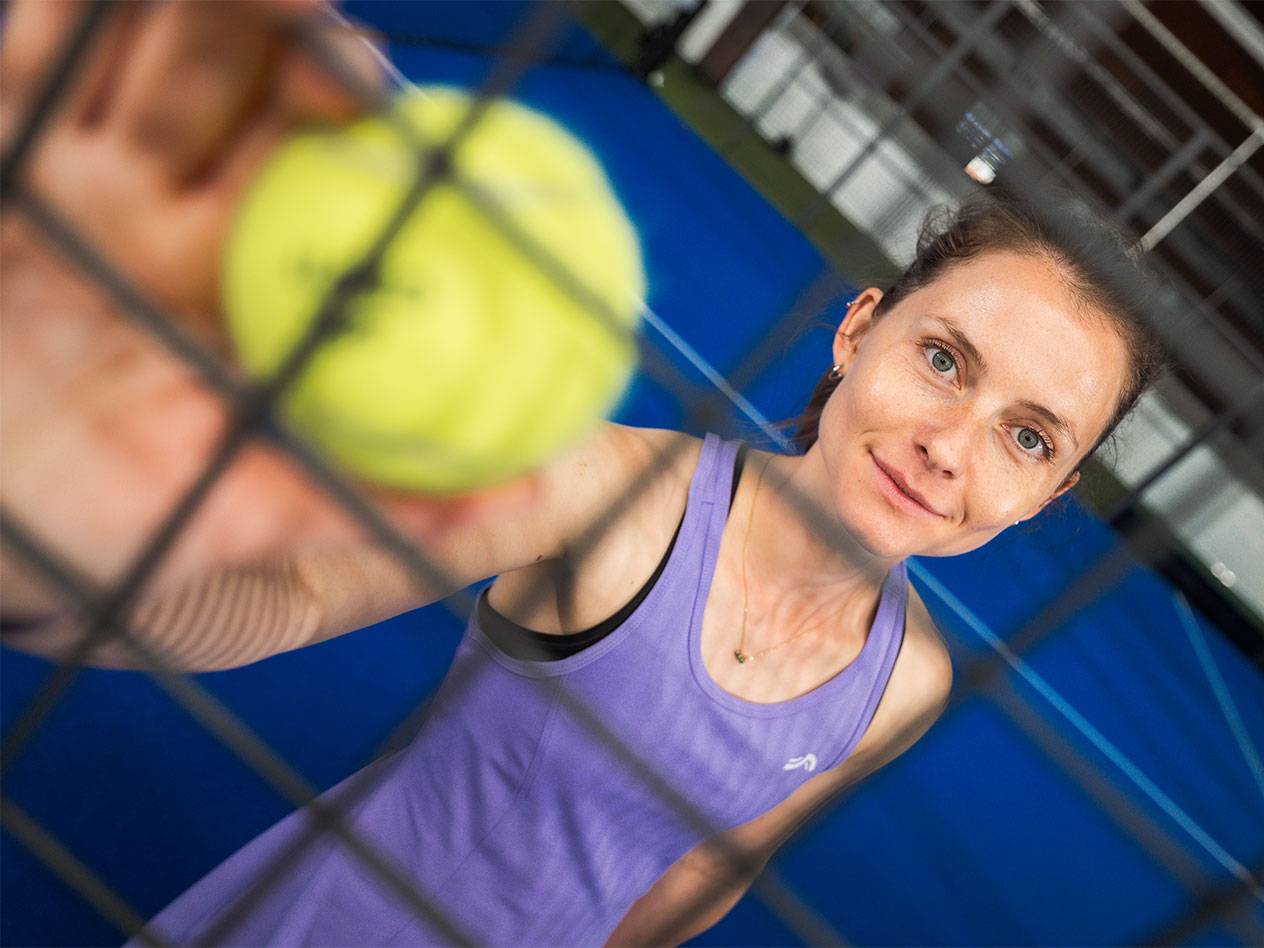 Lana Büttner in sportswear holds a padel ball through a net, with a blue court in the background.