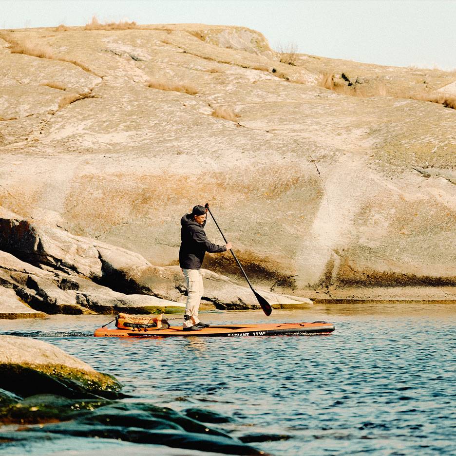 Man paddling an orange stand-up paddleboard on a rocky lake.