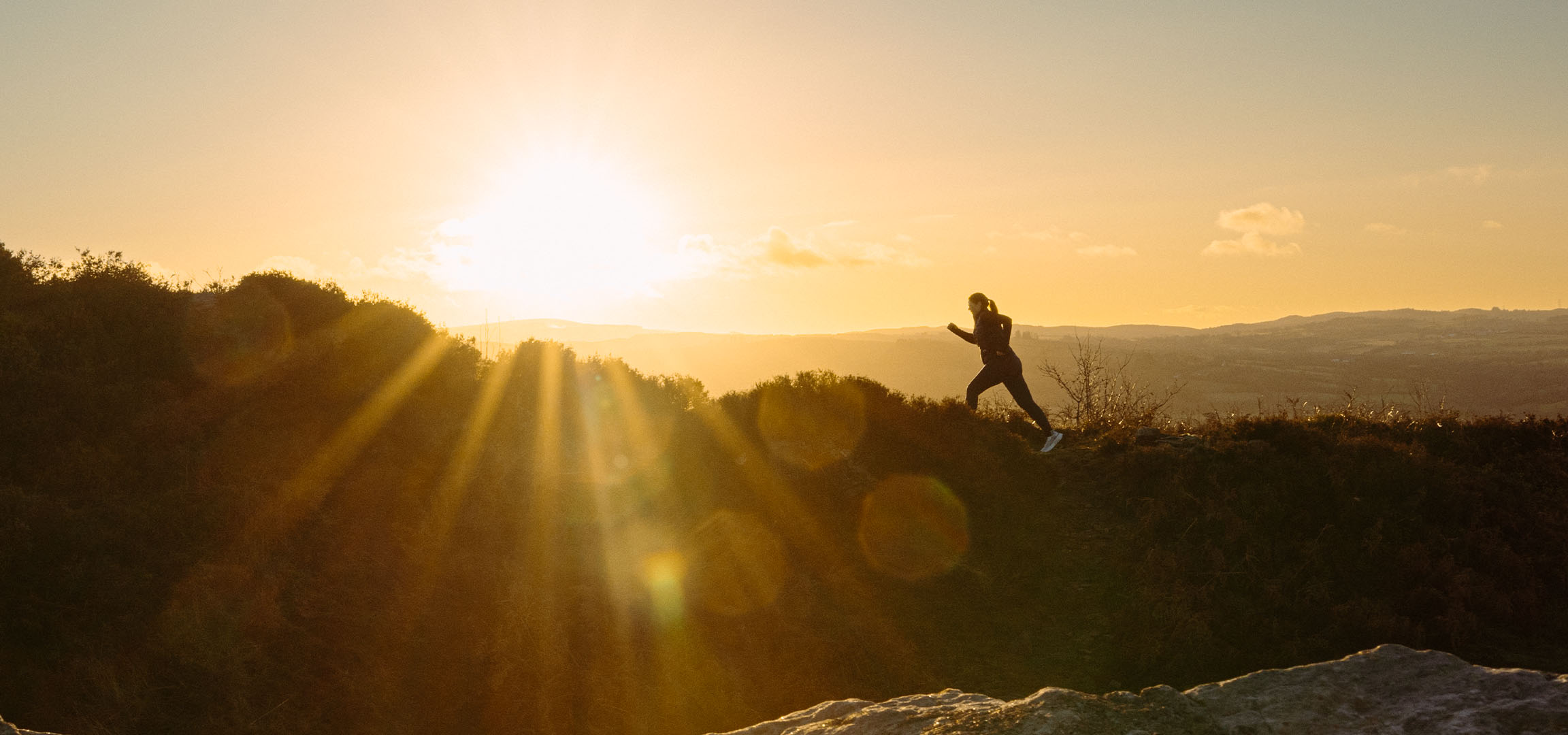 Woman running on a hill at sunset, with sun rays and a scenic landscape.