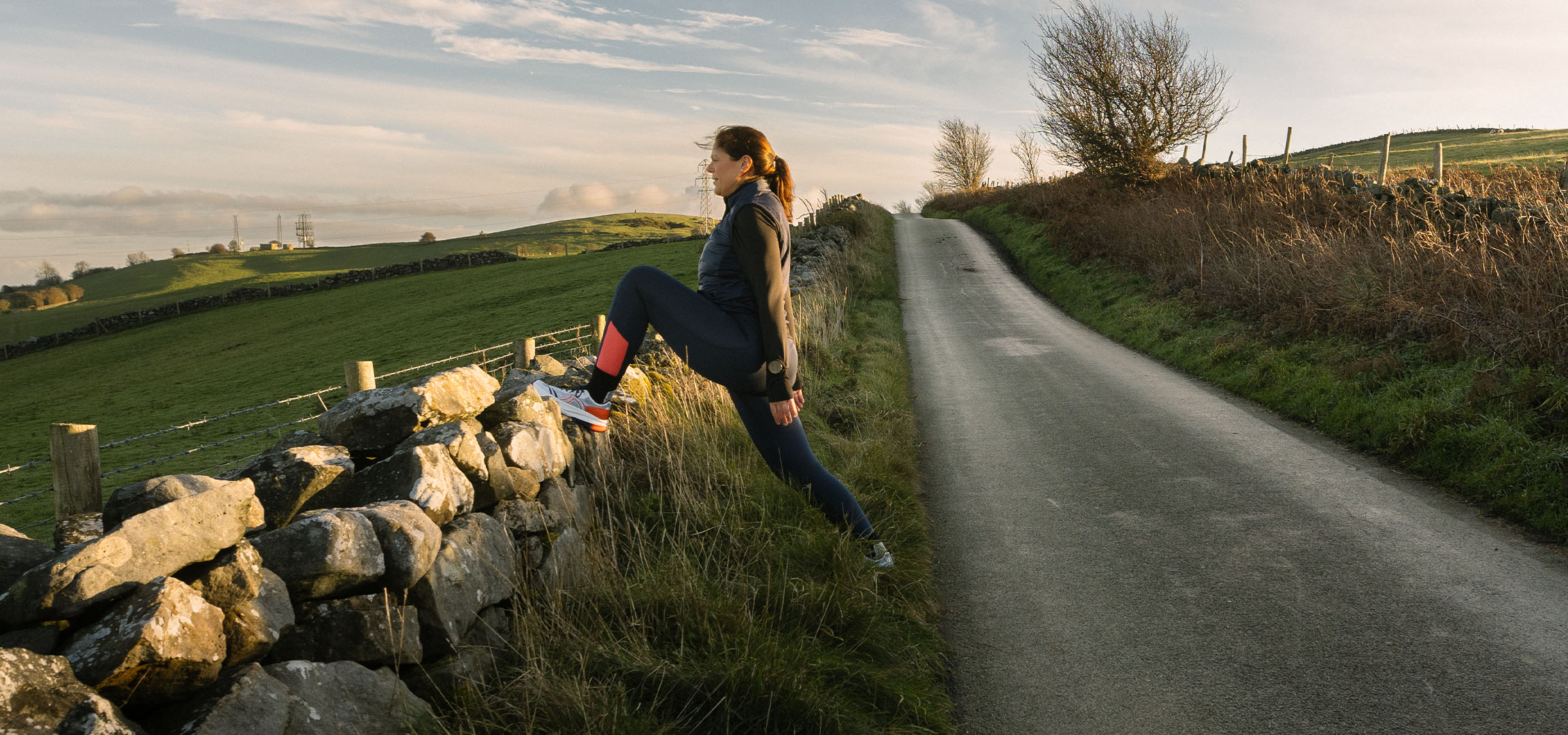 Woman stretching by a stone wall on a country road at sunset.
