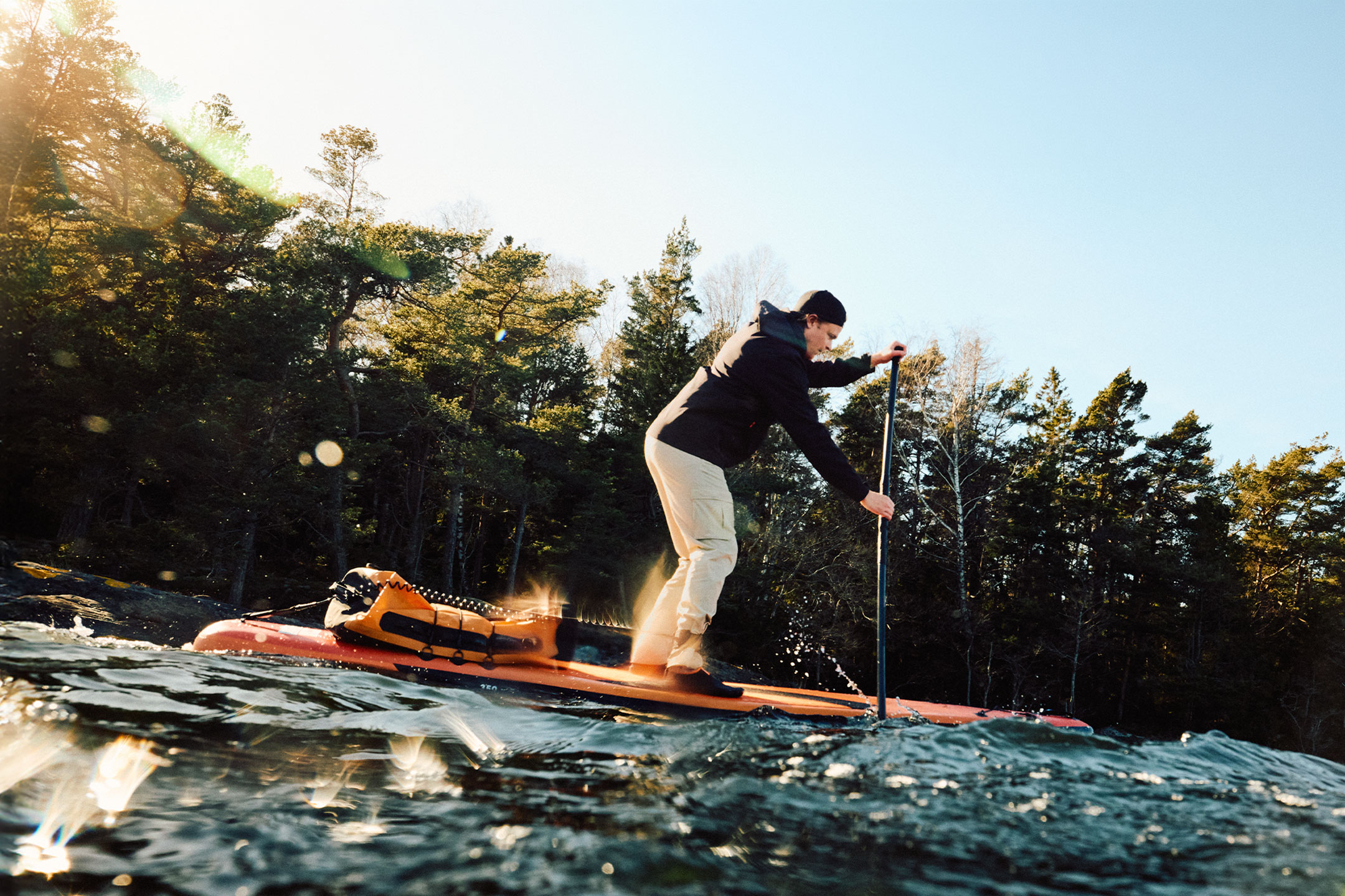 Man paddleboarding with an orange dry bag next to a forest.
