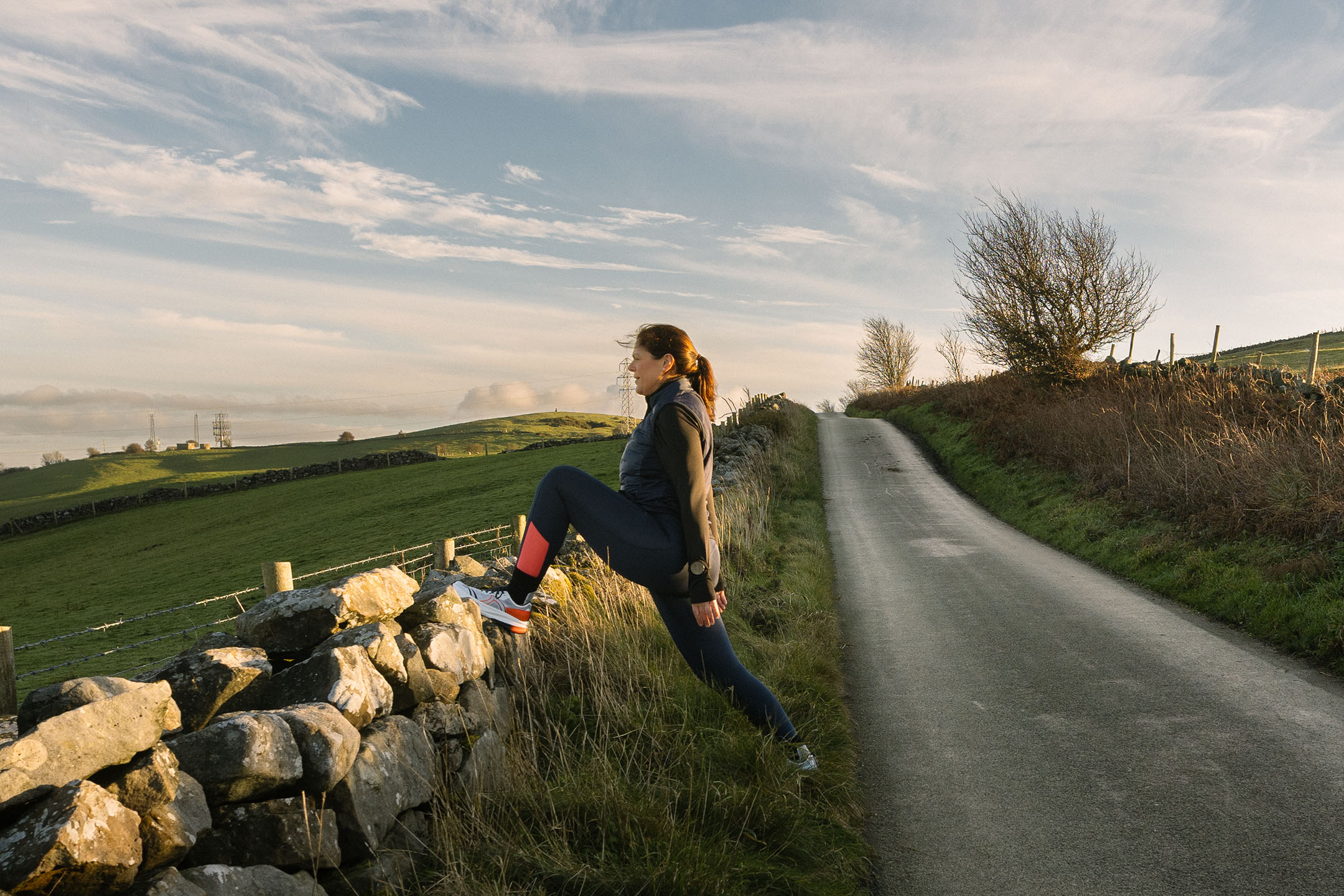 Woman in sportswear stretching on a stone wall beside a rural road.