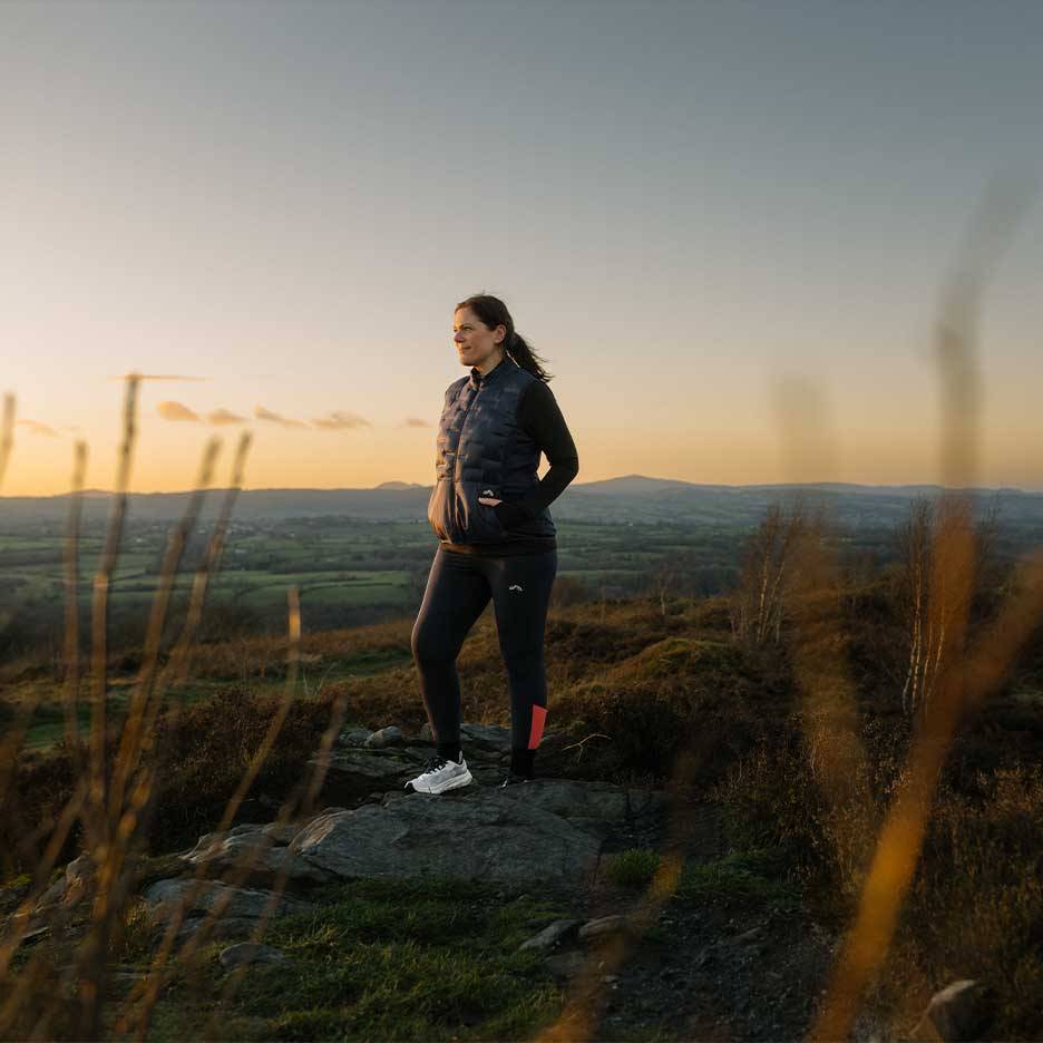 Woman in a quilted vest and leggings stands on a hilltop at sunset.