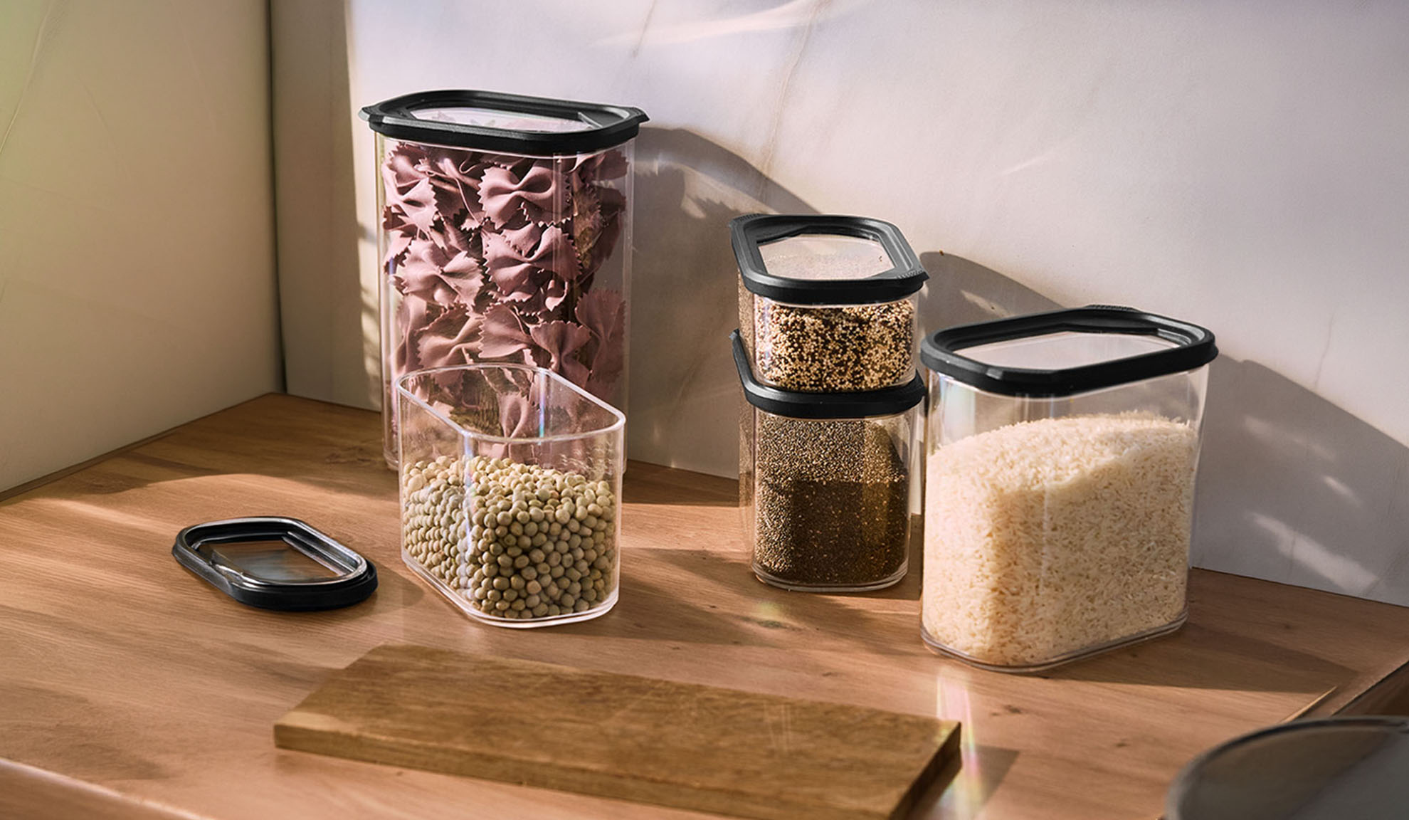 Clear kitchen storage containers with pasta, lentils, seeds, and rice on a wooden countertop.
