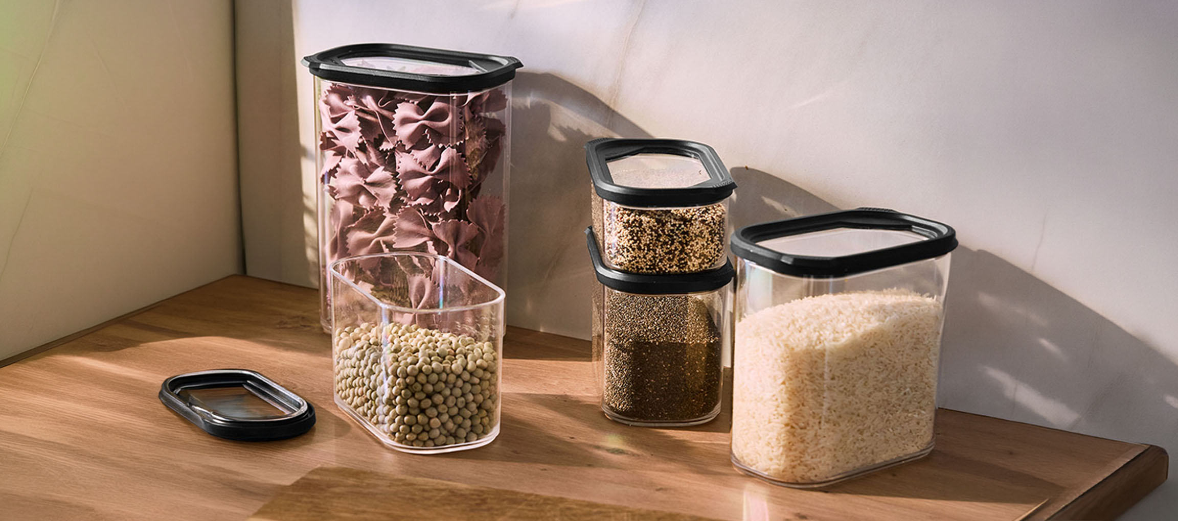 Clear kitchen containers with black lids, filled with pasta, lentils, quinoa, chia seeds, and rice on a wooden surface.