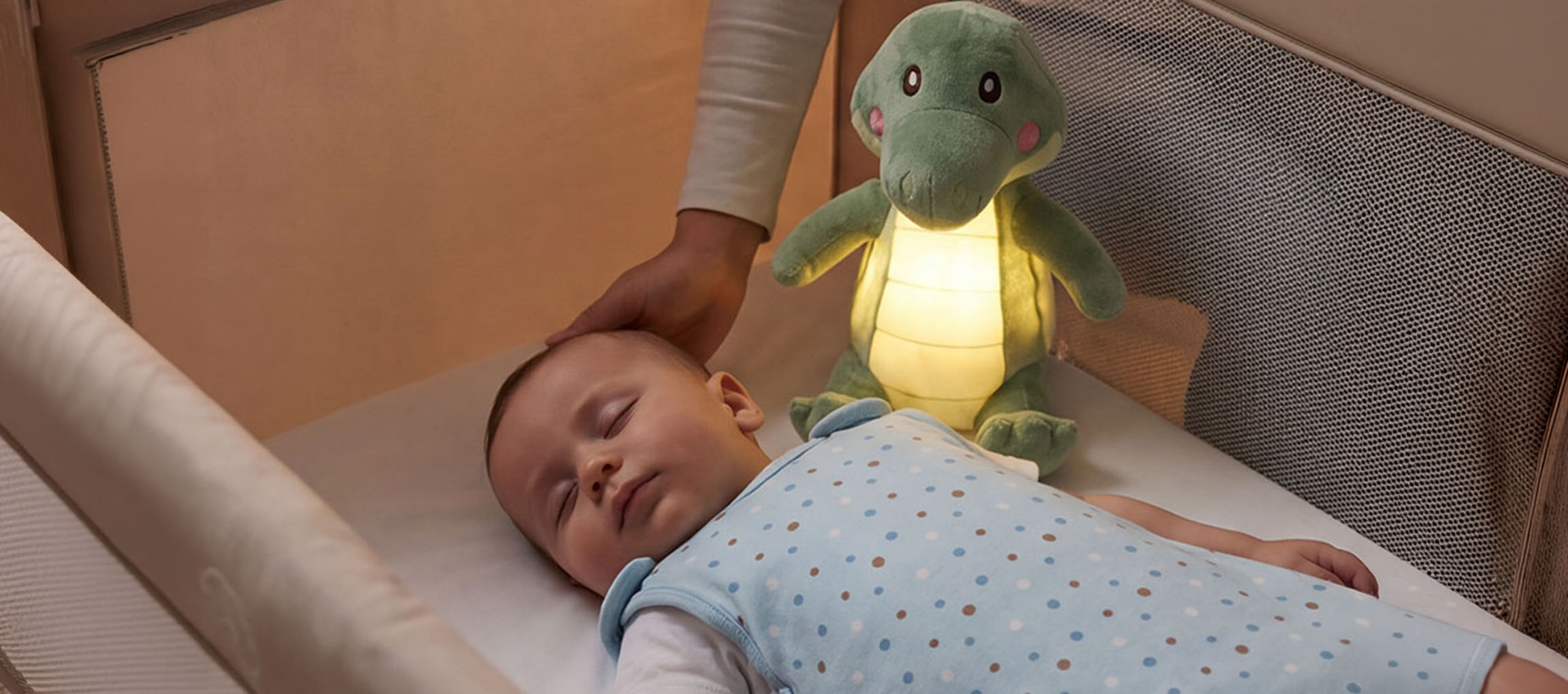 Baby sleeping in a crib with a plush night light, adult stroking the baby's head.