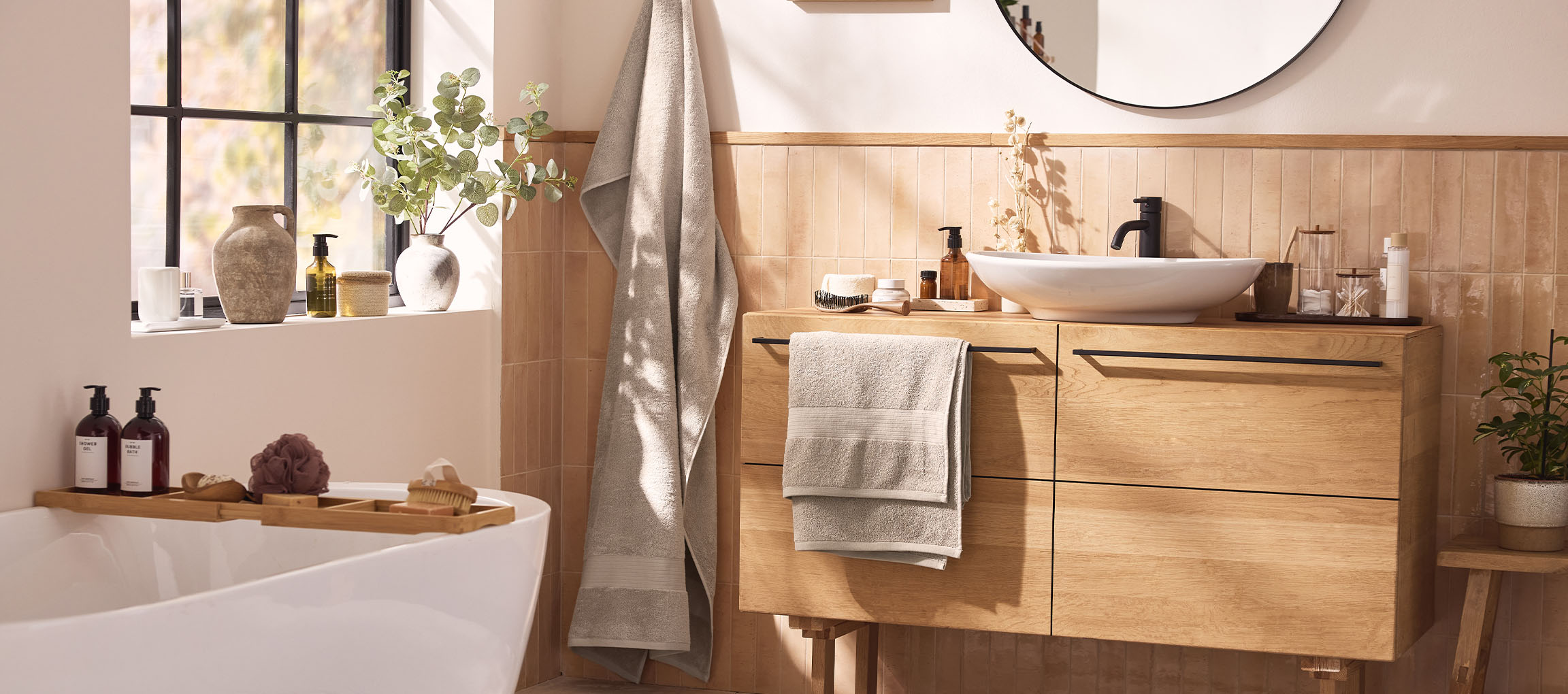 A modern bathroom with a wooden vanity, white sink, round mirror, and bathtub with bath accessories.