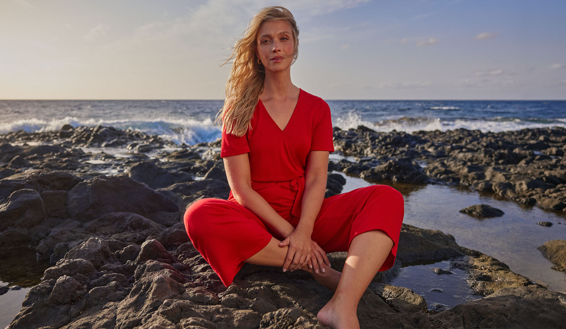 Woman in red jumpsuit sitting on rocks by the sea.