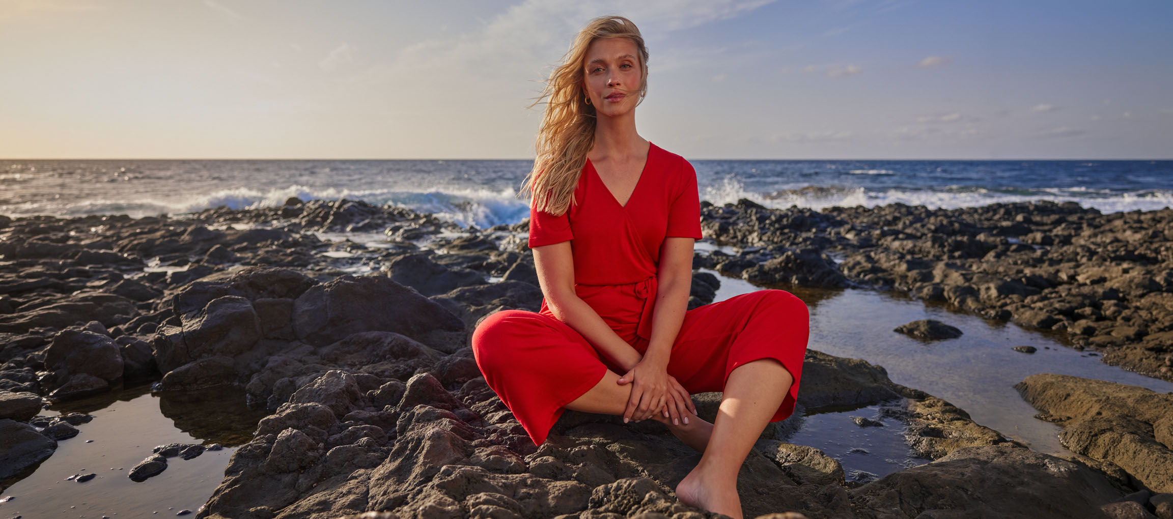 Woman in a red jumpsuit sits on a rocky beach by the sea.