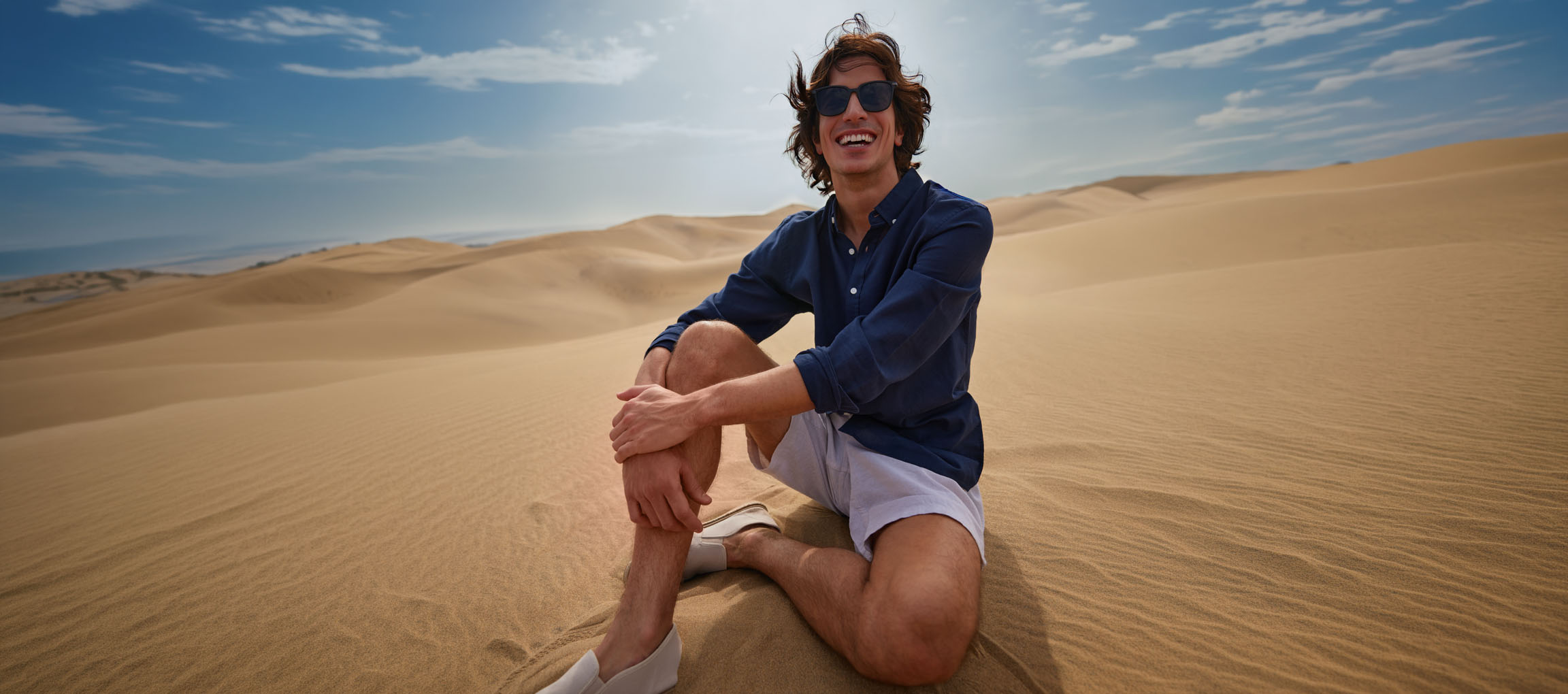 Man in a dark blue shirt and shorts sits in sand dunes, smiling.