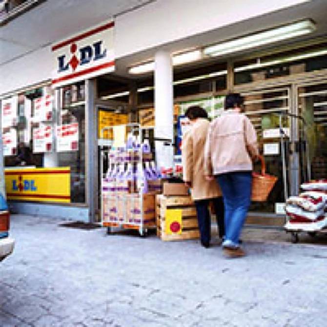 Two shoppers enter a Lidl store with a basket, surrounded by various products on display.