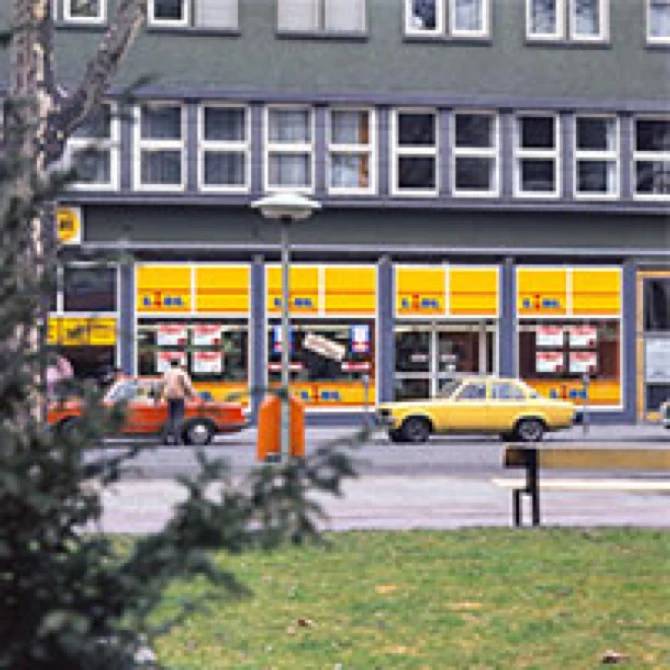 A Lidl store with yellow window displays, cars parked outside, and a building above.