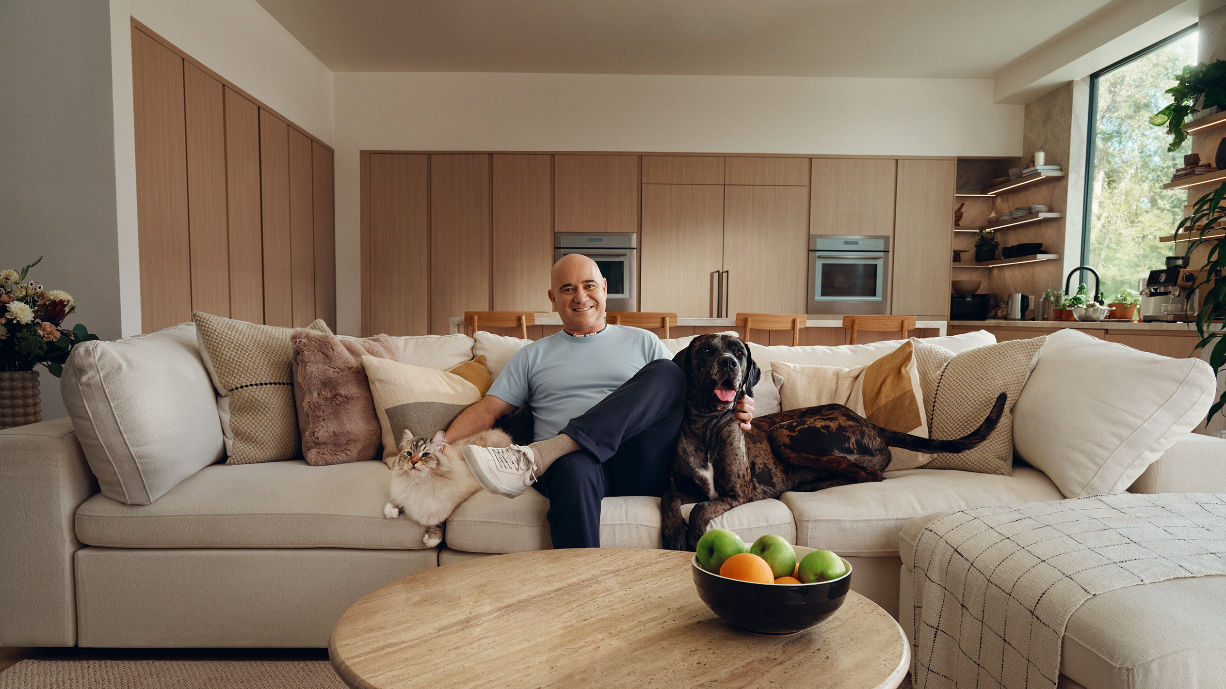 Andre Agassi sits on a couch with a cat and dog, with a fruit bowl on the coffee table.